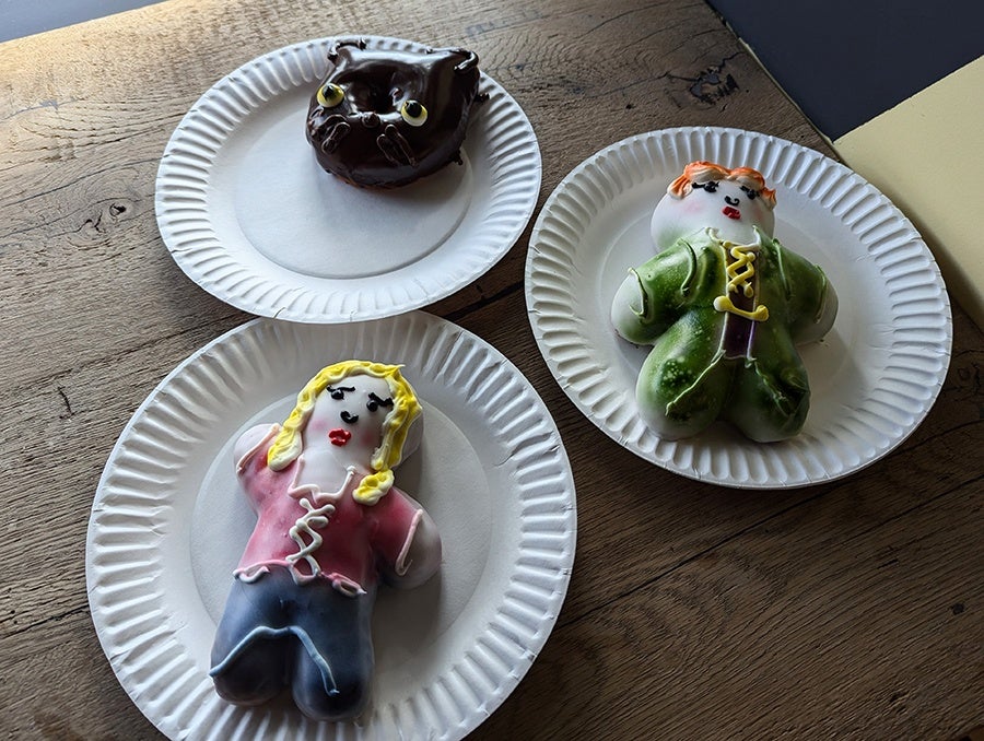 Three decorated doughnuts—two witches from the movie Hocus Pocus and a black cat—sit on paper plates on a wooden counter.