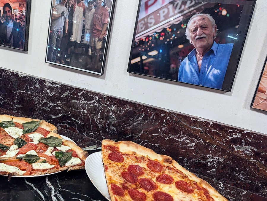 Two pepperoni slices and two basil-topped slices with fresh tomato sit on a counter under a photograph of a pizzeria owner.