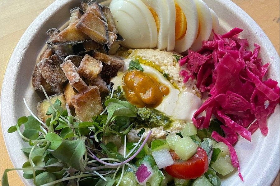 Overhead view of a bowl of fried eggplant, hummus, pickled onions, hard boiled eggs, cucumbers, and more.