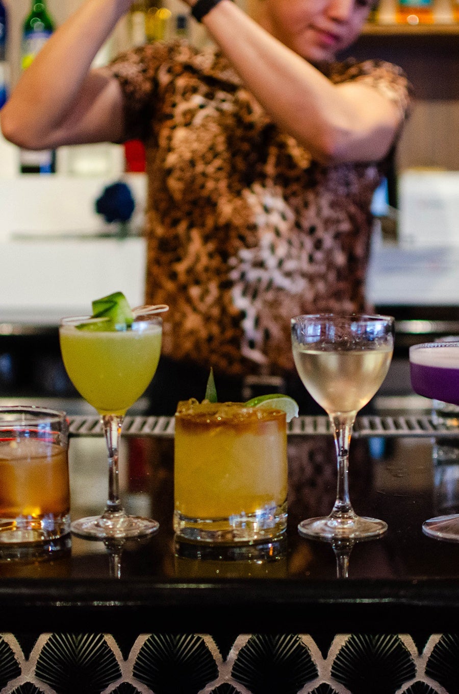 A bartender in a leopard-print shirt shakes a cocktail behind a lineup of cocktails on a bar.