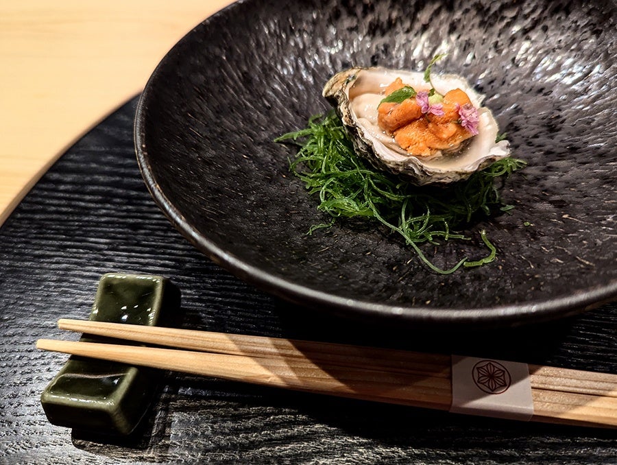 An elegant uni-topped oyster sits on seaweed on a black plate.