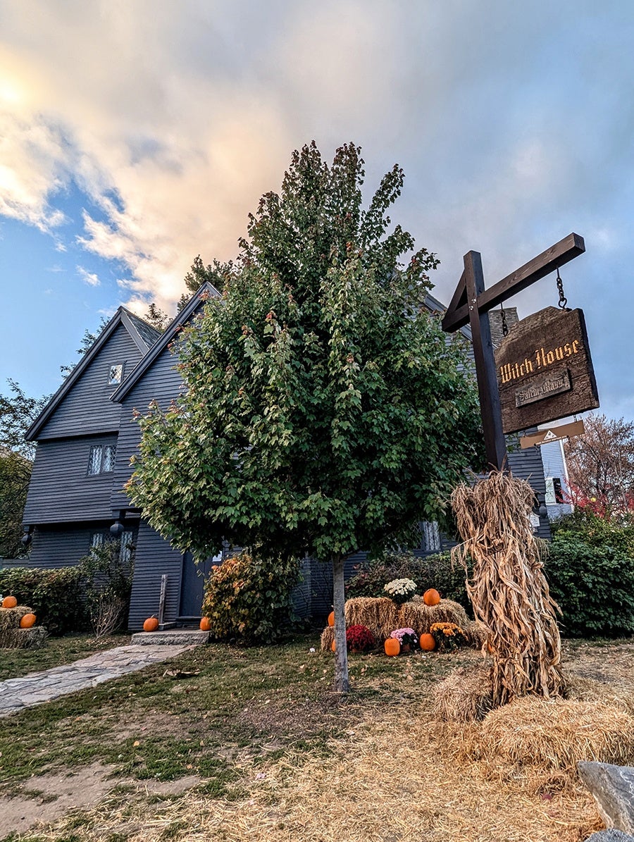 A historical black house with a sign in front that says Witch House is decorated with pumpkins.