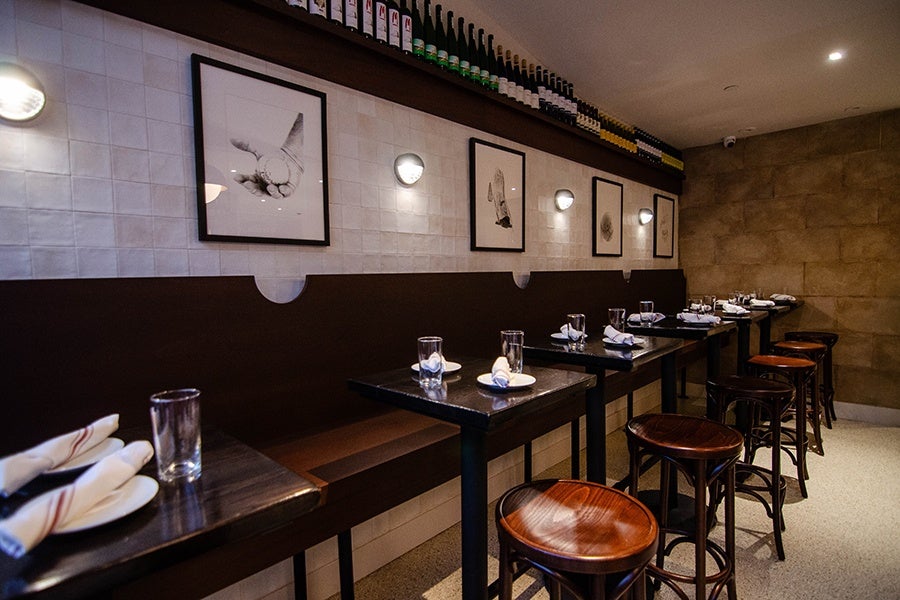 A dining room of a restaurant, with white tiles behind the banquette and glossy wooden stools.