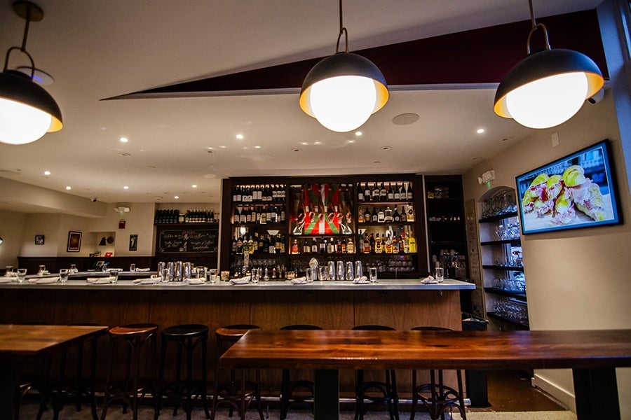 The bar area of a Basque-inspired pintxo bar, with a couple standing tables in front and some legs of jamón hanging in view behind the bar.