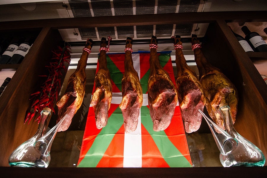 Legs of jamón hang behind a bar near two porróns and a Basque flag.