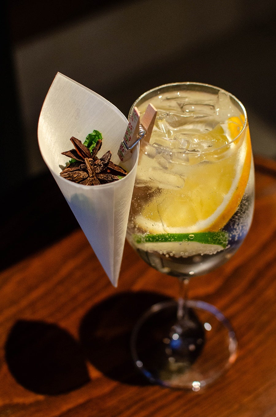 A gin and tonic has citrus slices in it and a paper cone full of herbs and spices clipped to the glass with a tiny clothespin.