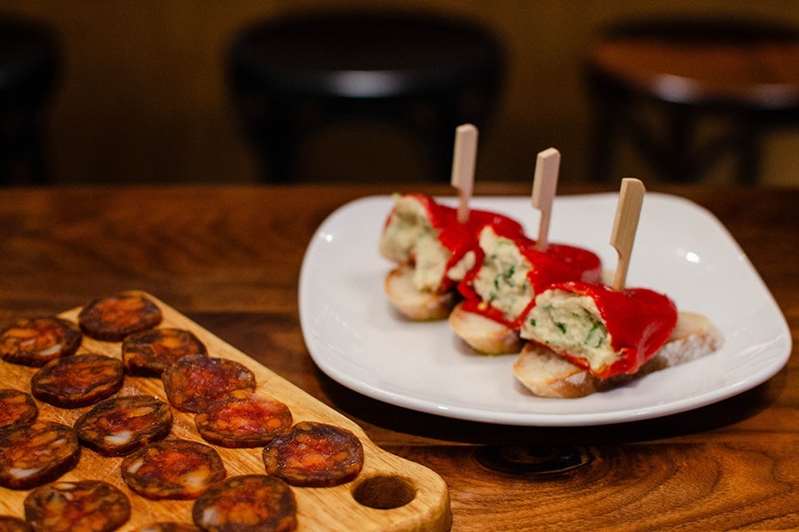 Long red peppers are stuffed with a thick salt cod filling with herbs, sitting next to coins of spicy sausage on a wooden board.