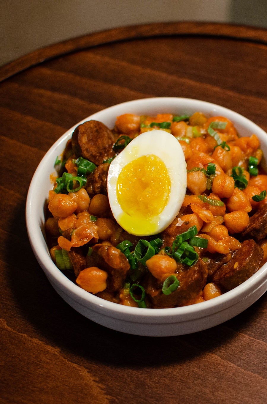 A bowl is full of chickpeas, sliced sausage, and half a boiled egg with a runny yolk.
