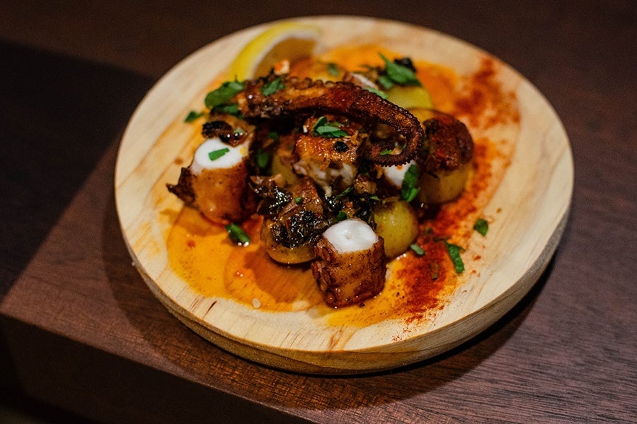 Charred octopus and little potatoes sit on a light wooden plate in a reddish oil.