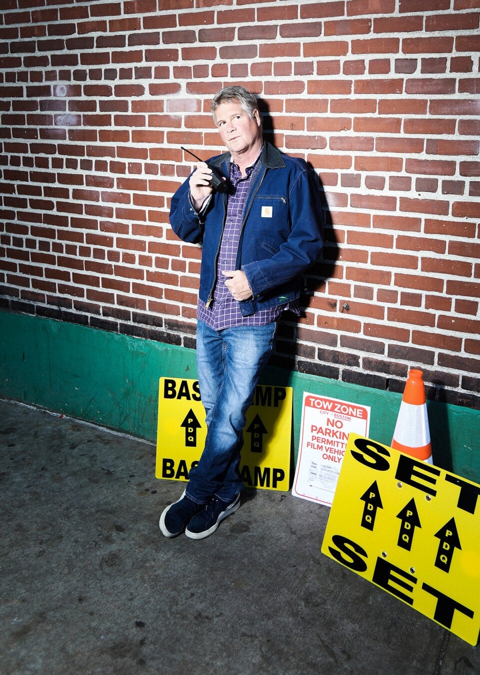 Man with gray hair standing against a brick wall with a denim Carhartt jacket and a walkie talkie.