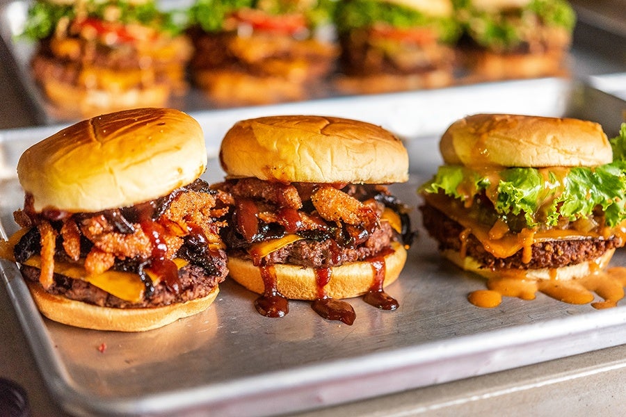 Lineup of three burgers on a metal tray with various toppings.