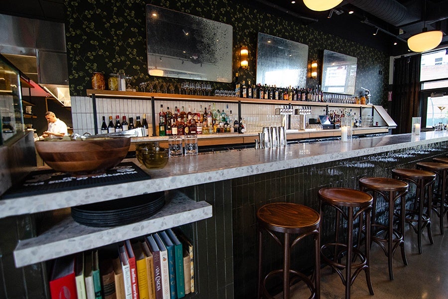 An restaurant bar features wooden stools, vertical dark green tiling, floral wallpaper, and large distressed mirrors.