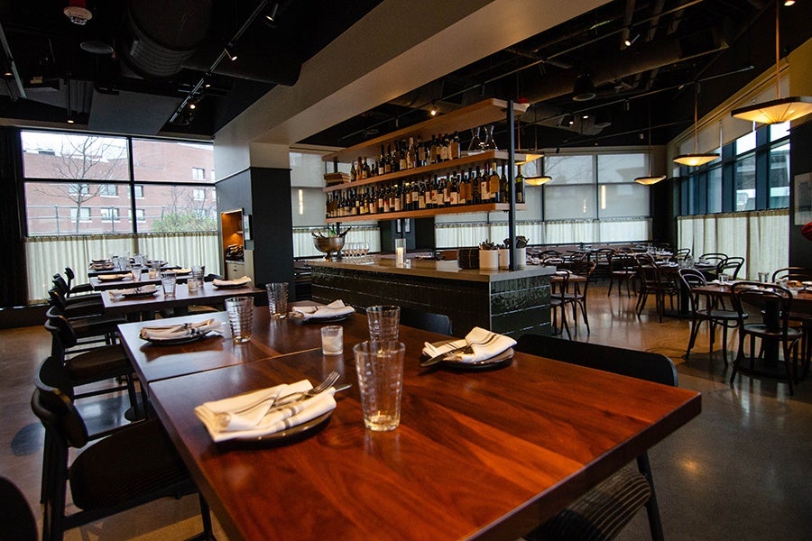 A restaurant dining room features long cherry wood tables and a central service station with shelves of wine bottles.