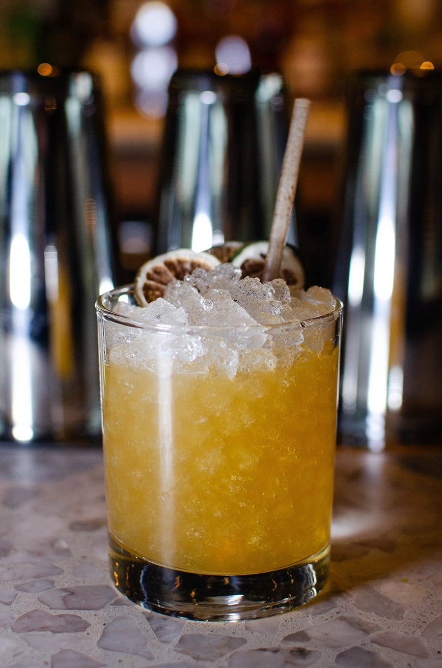 An orange cocktail over lots of crushed ice sits in front of silver shakers.