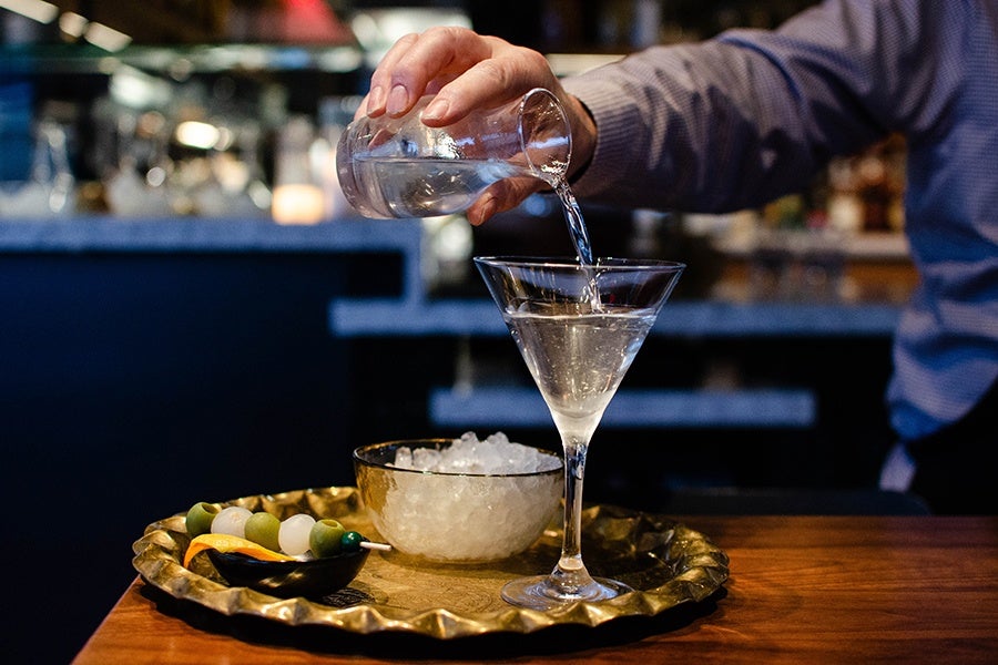 A hand pours a martini from a carafe into a glass on an elegant tray.
