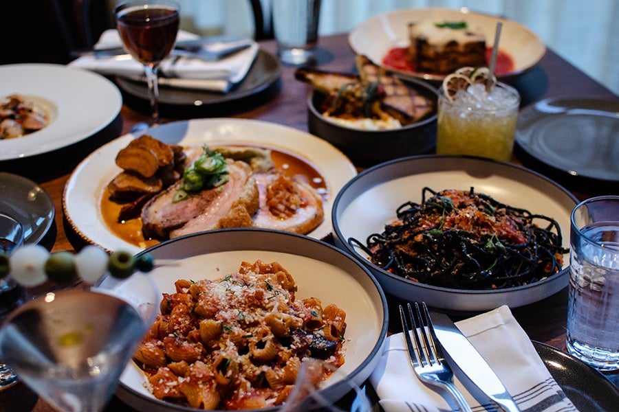 A spread of pasta dishes, porchetta, cocktails, and more on a dark wooden table.
