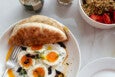Overhead view of eggs in a pool of labneh and olive oil with a pita, plus a yogurt parfait in a nearby bowl.