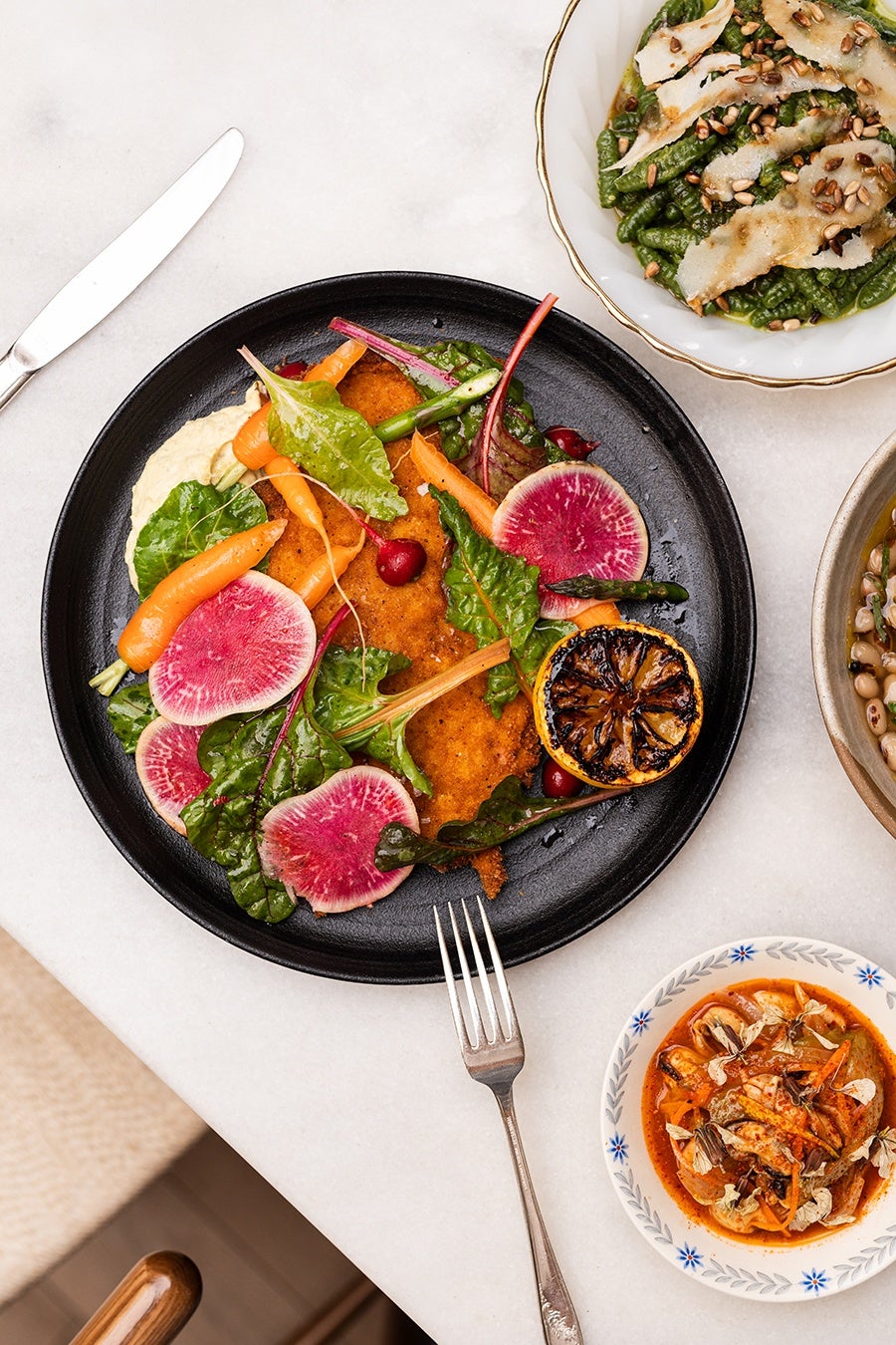 Overhead view of a few dishes at a restaurant, including a thick green pasta and a crispy cutlet topped with sliced radishes and little carrots.