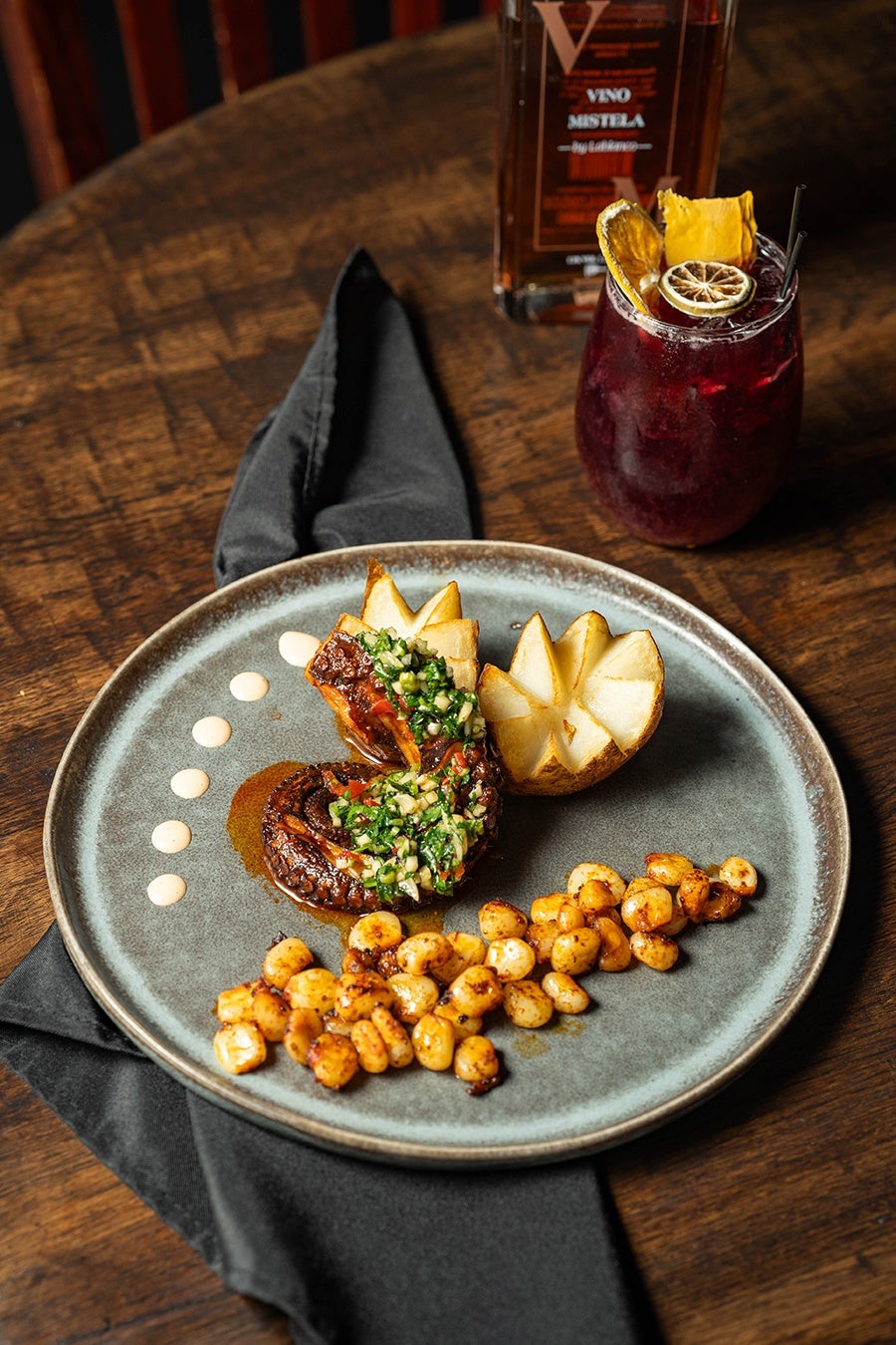 Grilled octopus tentacle is plated with dollops of a creamy sauce and an herby topping. A cocktail sits to the side of the plate.