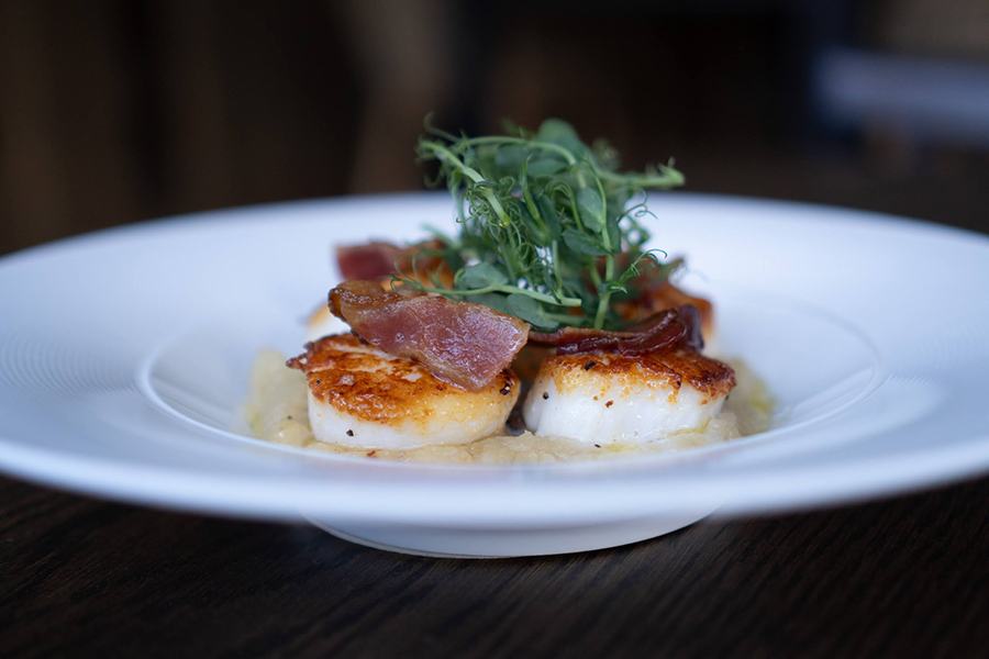 Seared scallops sit in a shallow bowl, topped with bacon and greens.