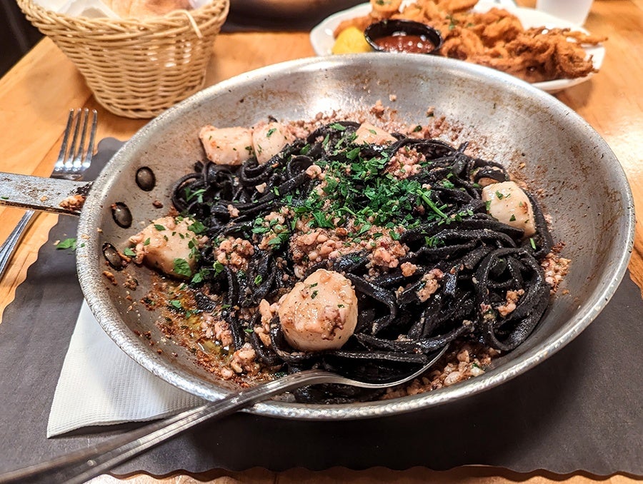 A silver pan full of black pasta and scallops, tossed with a ground calamari and herb topping.
