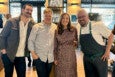 Four smiling people stand in a line in a restaurant, looking at the camera. One is in a chef's apron.