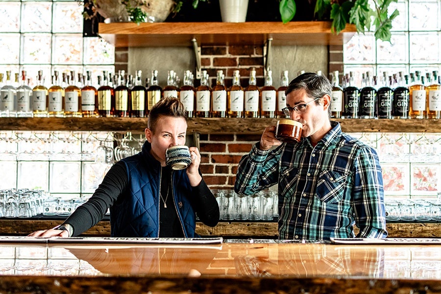 Two people stand behind a bar, sipping from coffee mugs.