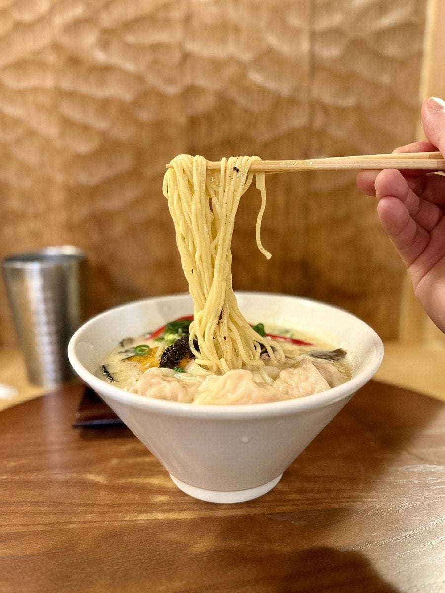 A hand pulls thin noodles up from a bowl full of soup.