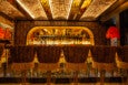 A bar features tasseled, upholstered seats, a mirror-and-exposed-brick ceiling, a mirrored backbar, and patterned wallpaper.