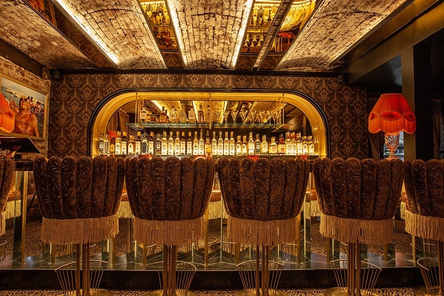 A bar features tasseled, upholstered seats, a mirror-and-exposed-brick ceiling, a mirrored backbar, and patterned wallpaper.