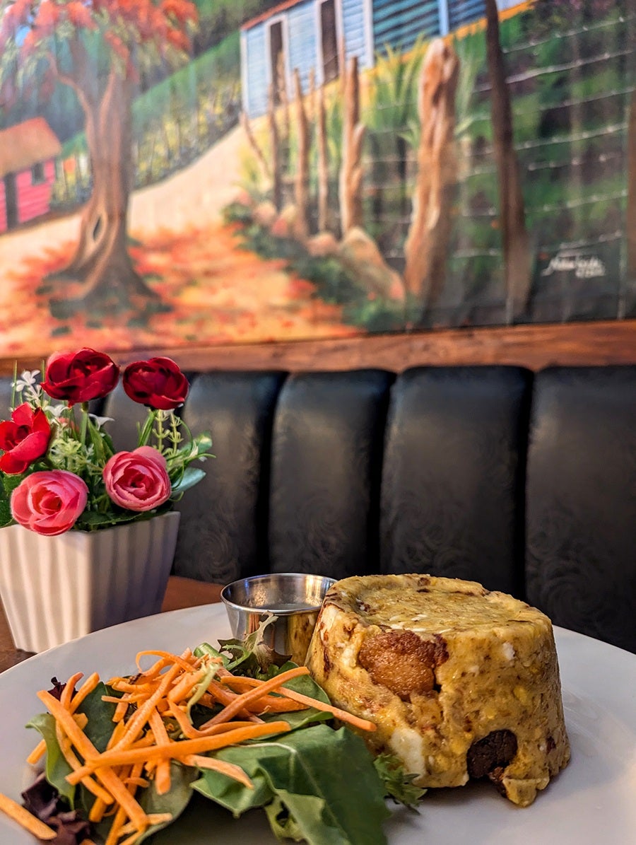 Mofongo sits on a plate with a side salad in a restaurant booth, with a colorful mural in the background.