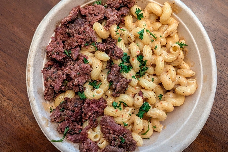 Overhead view of a takeout bowl of cavatappi and cheese with a crumble of lamb sausage and an herb garnish.
