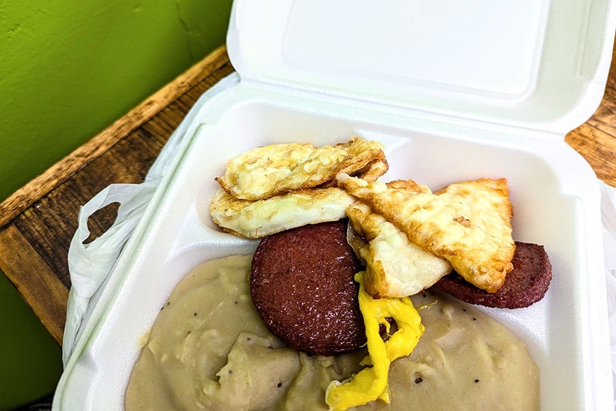A white styrofoam takeout container is filled with smooth plantain mash, coins of salami, fried cheese, and eggs.