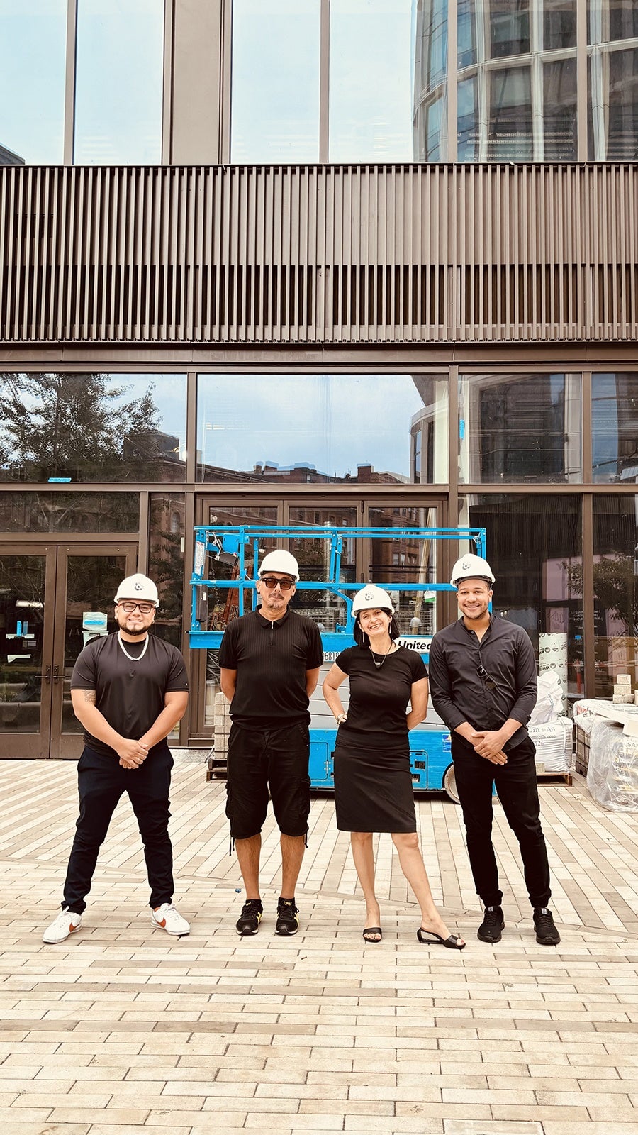 Four people dressed in black clothes and white hard hats smile, lined up in front of an under-construction space in a tall building.