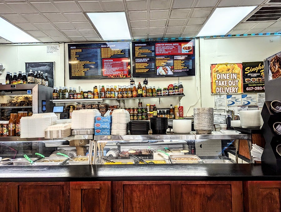 The counter at a casual Trinidadian restaurant. The menu on the wall features roti, buss up shut, cocoa bread sandwiches, and more.