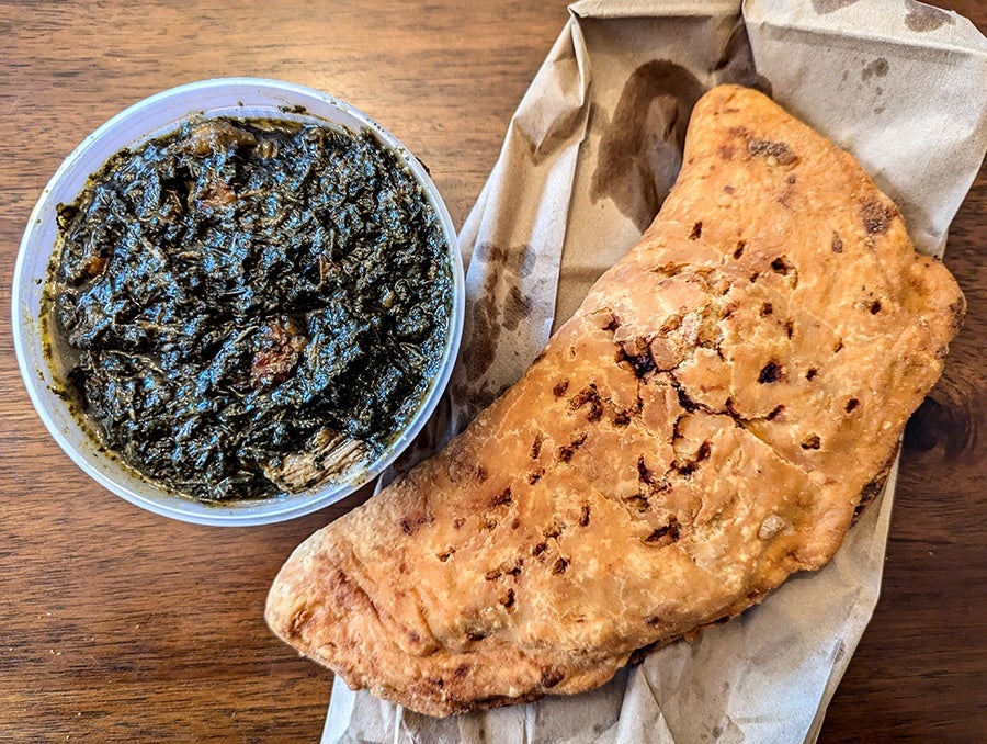 Overhead view of a takeout container of a dark stewed leafy green next to a giant doughy Haitian patty.