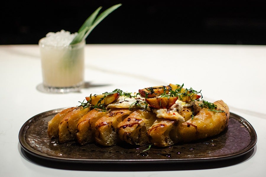 A small loaf of focaccia is sliced and topped with cheese, peaches, basil, and drizzles of balsamic and olive oil, next to a yellow cocktail garnished with pineapple fronds.