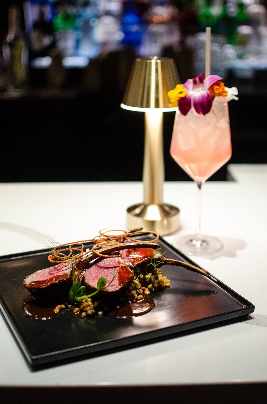 Rare lamb sits atop fregola on a black square plate on a white marble bar, next to a pink cocktail garnished with flowers.