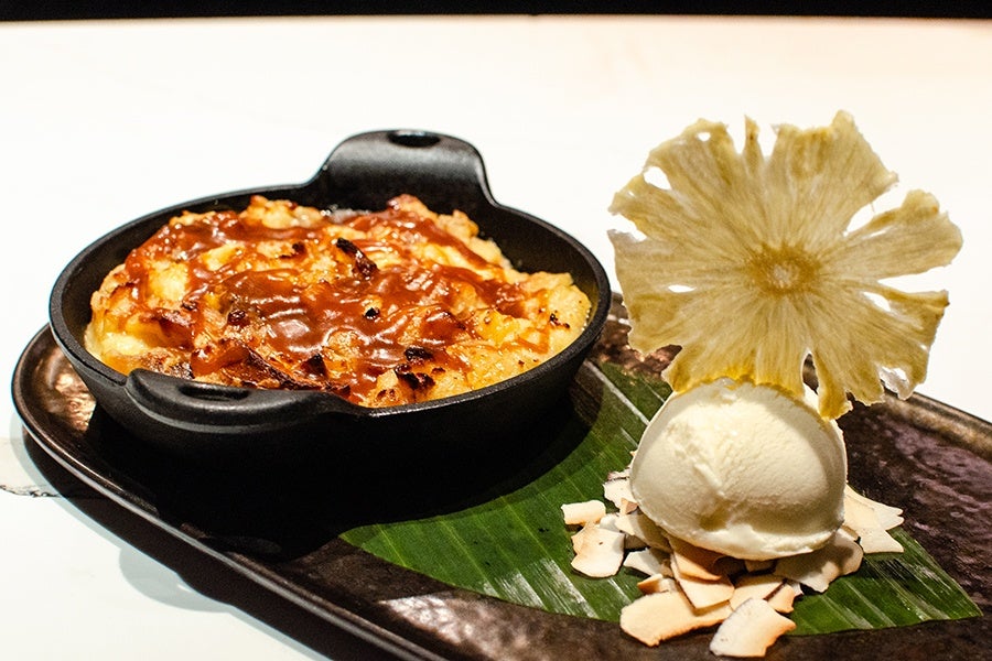 A bread pudding is drizzled with caramel and served in a small cast iron pan next to a scoop of ice cream garnished with a dehydrated pineapple slice.