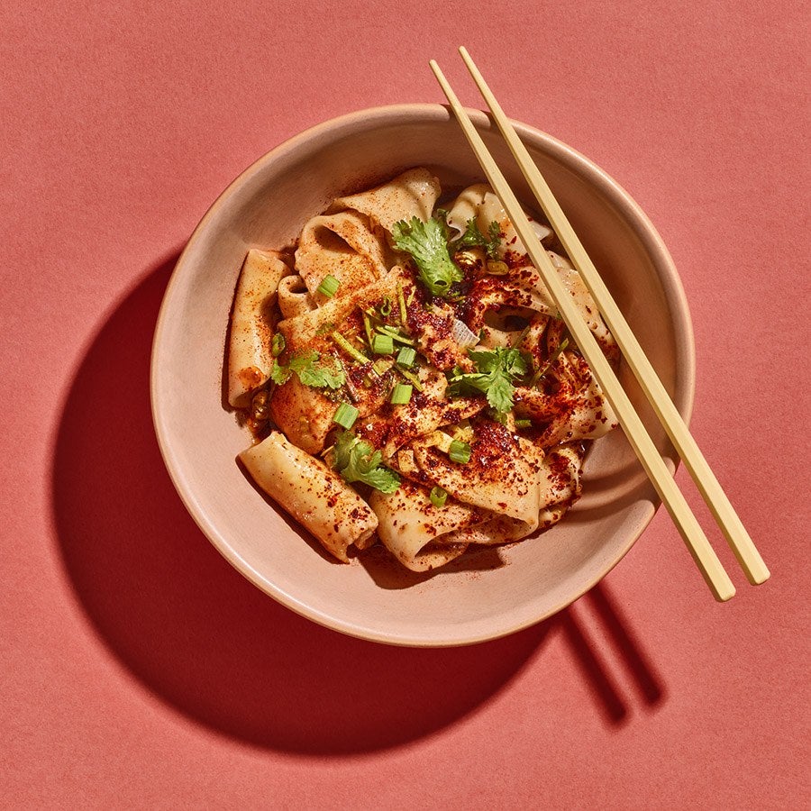 Overhead view of thick Xi'an-style hand-pulled noodles topped with chili powder and herbs. Chopsticks sit atop the bowl, which is on a pale red background.
