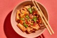 Overhead view of thick Xi'an-style hand-pulled noodles topped with chili powder and herbs. Chopsticks sit atop the bowl, which is on a pale red background.
