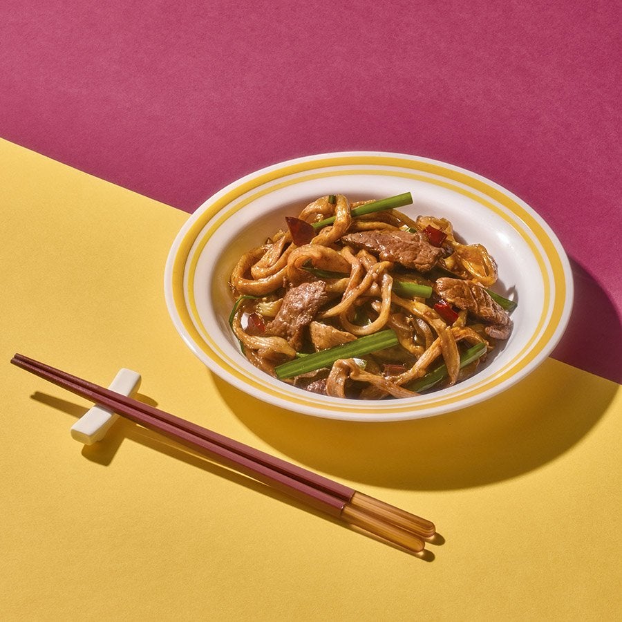 A bowl of noodles with slivers of beef and scallions sits on a red and yellow background next to chopsticks.