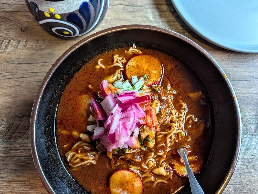 Overhead view of a bowl of ramen in a thin brown broth with sliced radishes, red onion, and other garnishes.