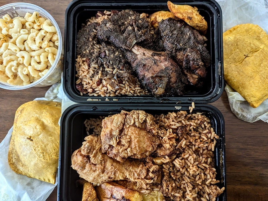 Overhead view of takeout containers of Jamaican food, including jerk chicken and fried chicken, each with plantains and rice.