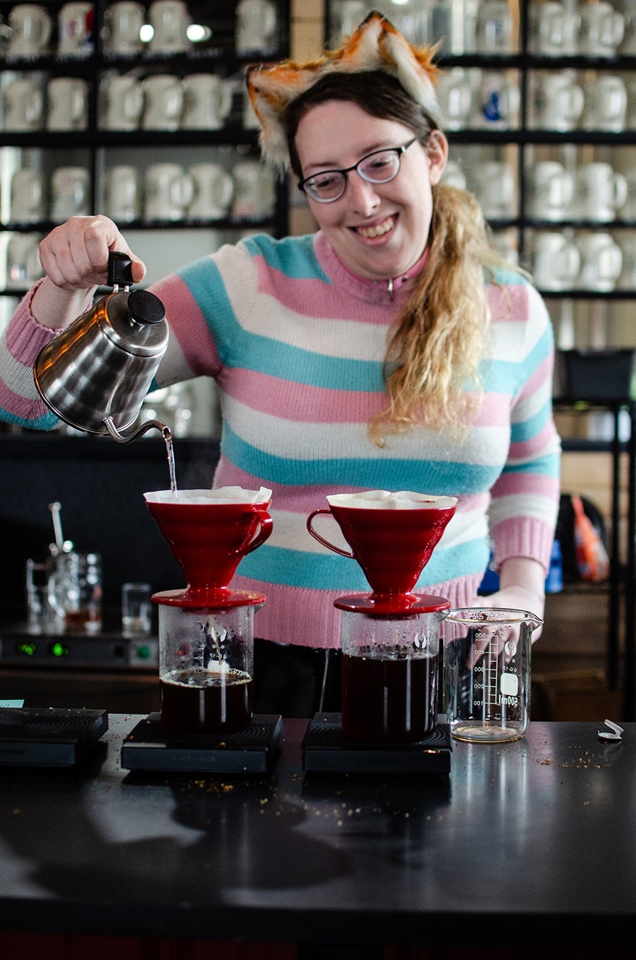 A smiling barista wearing fox ears and a blue, pink, and white striped sweater pours water into a pour-over coffee setup.