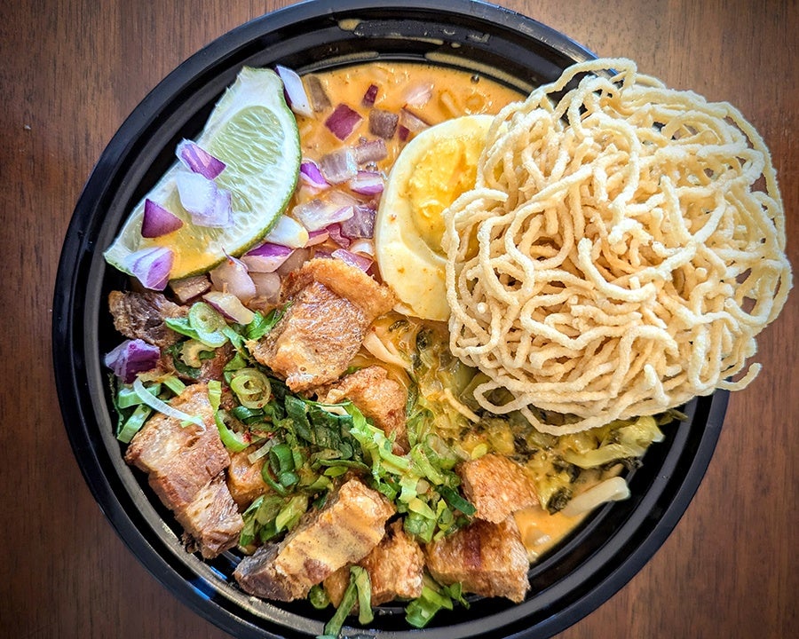 Overhead view of a takeout container of Thai khao soi, a pale yellow-orange curry with chunks of pork belly, mustard greens, lime, chopped red onion, and a bundle of crispy noodles.