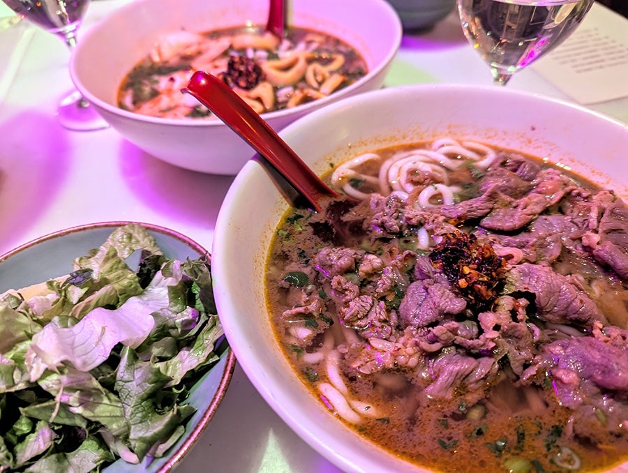 Two bowls of noodle soup, one with beef and one with seafood, sit on a table with glasses of sake.