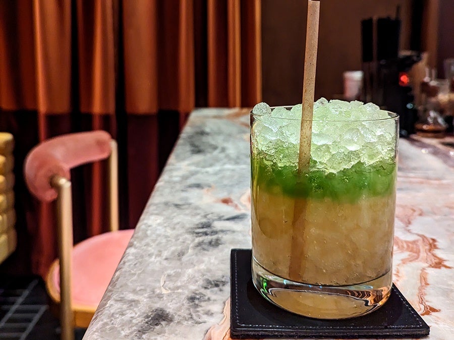 A drink, mostly pale yellow with a layer of bright green at the top, sits on a marble bar in front of a heavy red curtain.