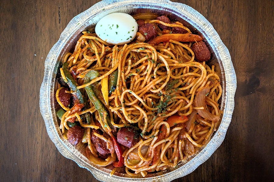 Overhead view of a takeout container of spaghetti with hard-boiled egg, hot dog bits, peppers, and onions.