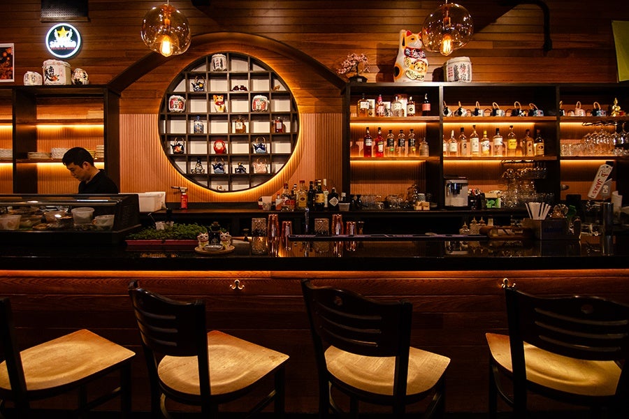 An employee of a restaurant stands behind the bar, which is decorated with Japanese teapots, waving cats, and other decor.